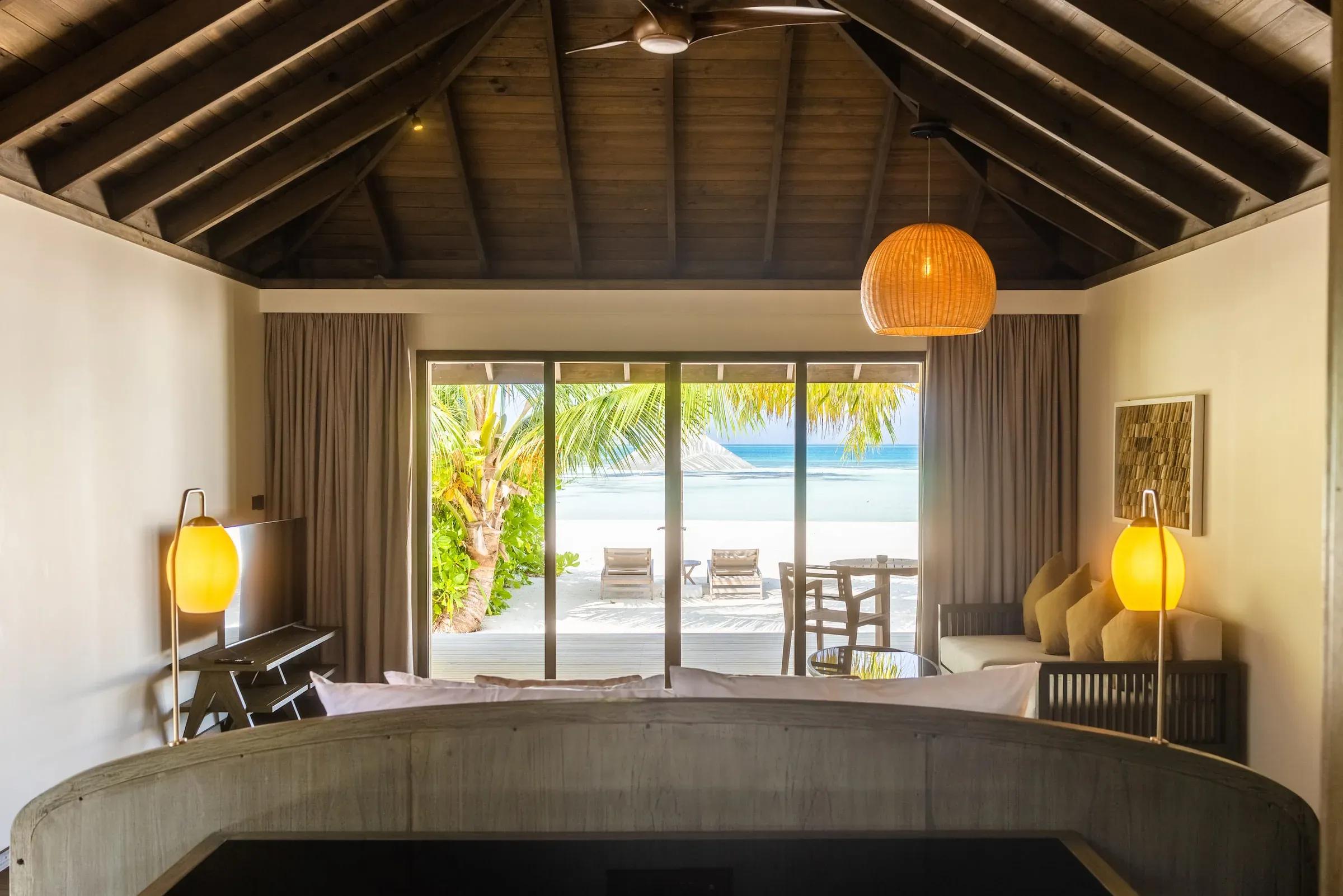 Interno della Dheru Beach Villa con vista su terrazza, lettini e mare al Jawakara Islands Maldives.